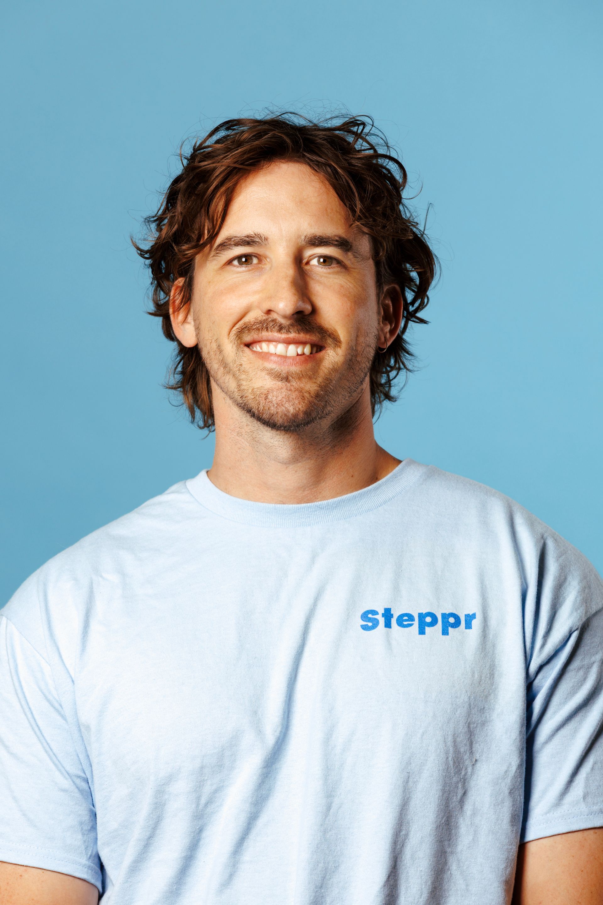 A smiling person with wavy brown hair wearing a light blue t-shirt with 