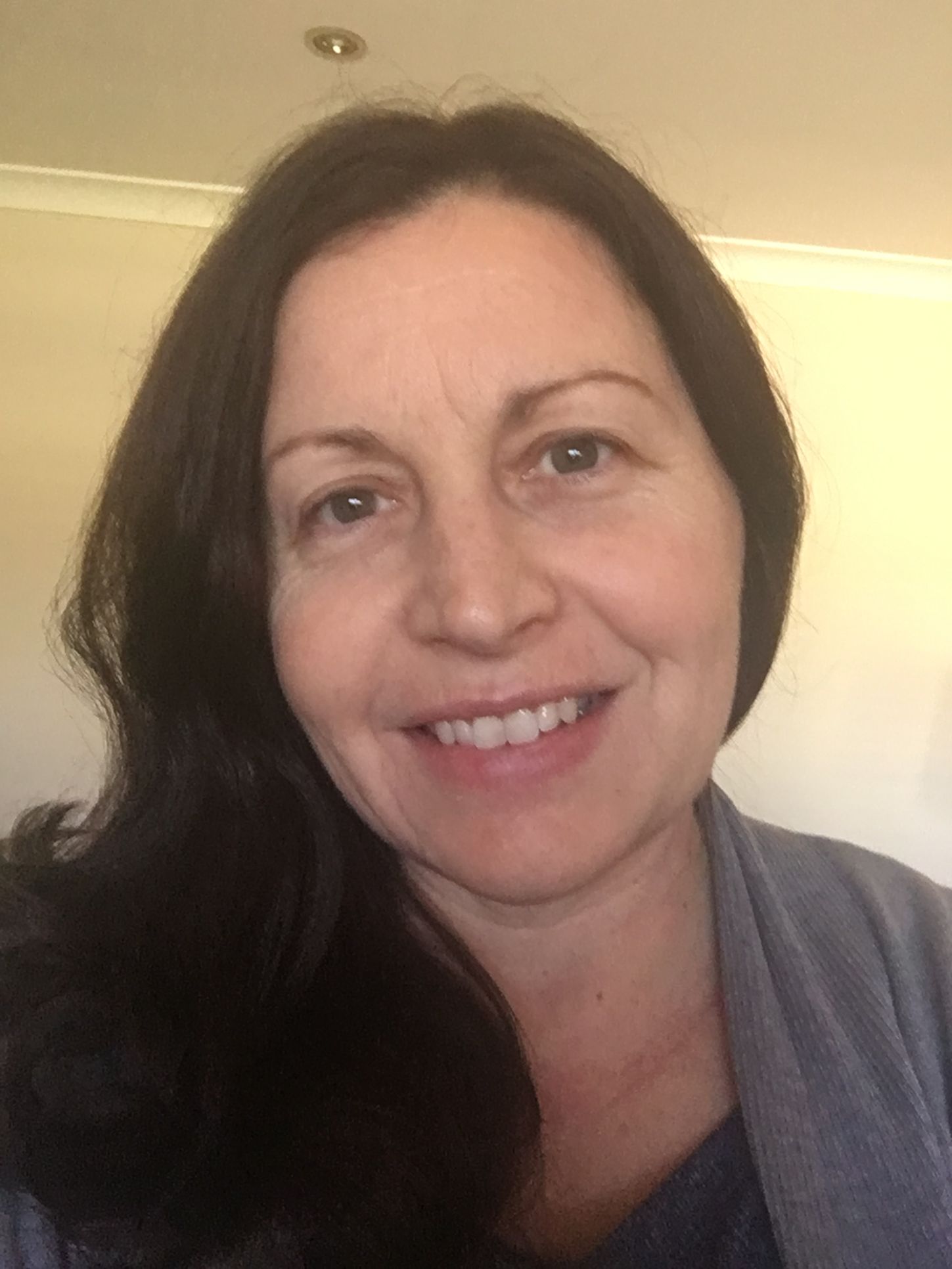 A smiling person with shoulder-length dark hair wearing a gray cardigan, captured in a close-up indoor portrait.