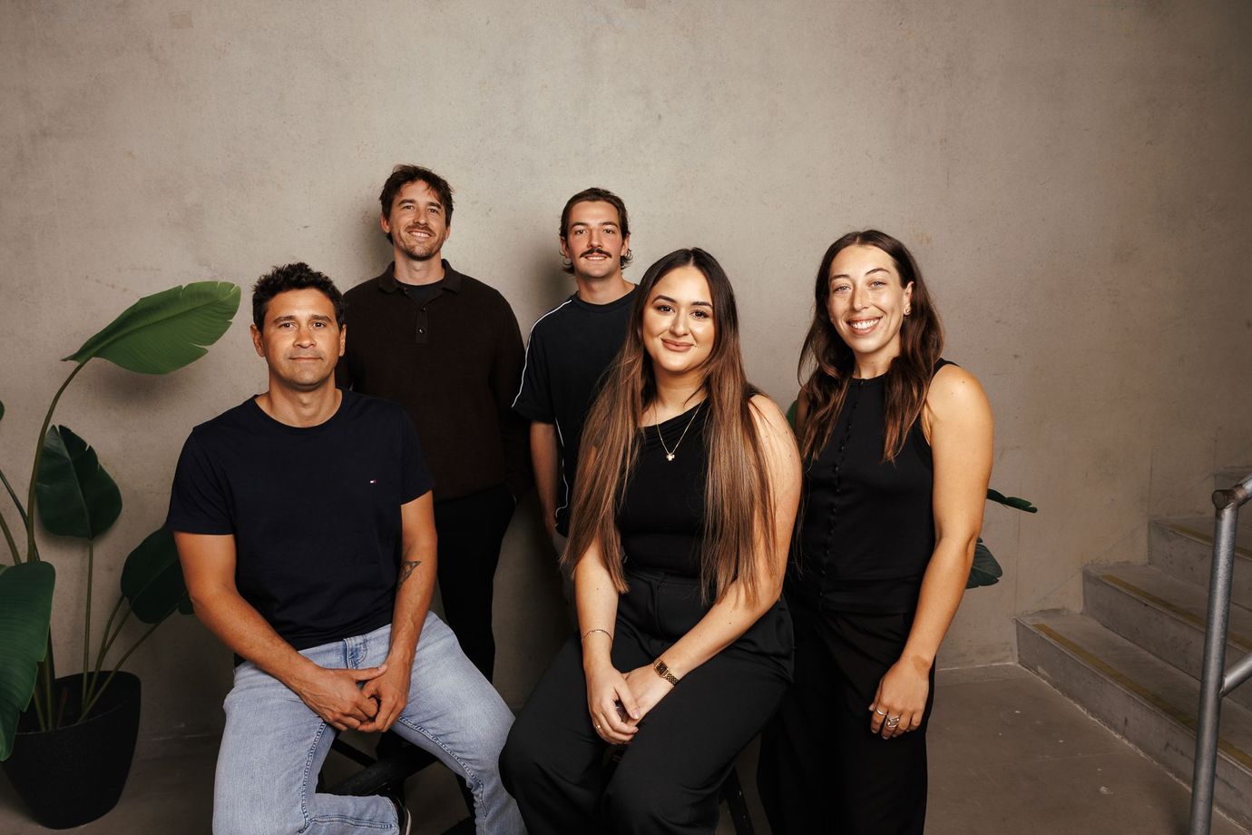 A diverse group of professional colleagues stand together in a bright, modern office, smiling toward the camera.