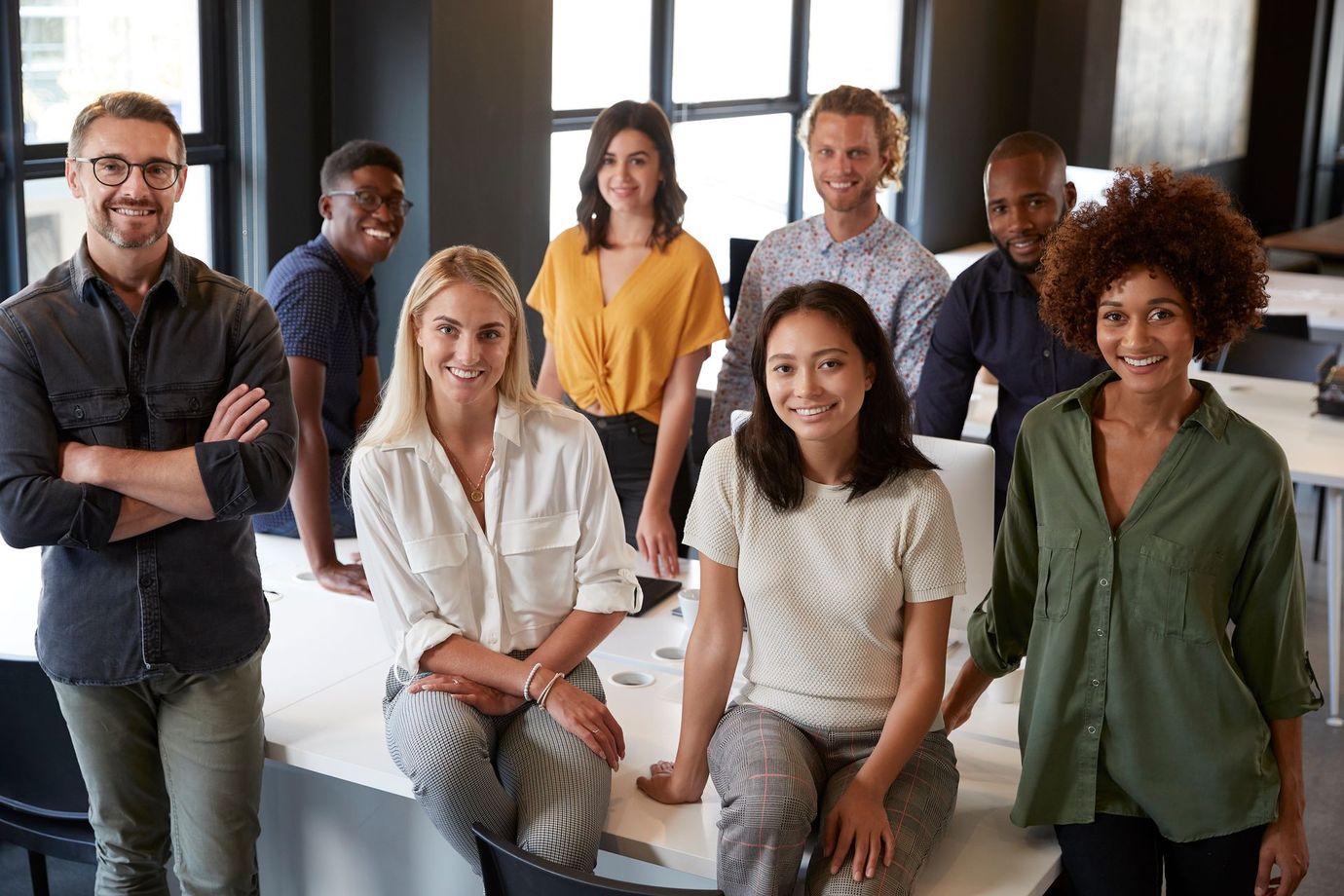 A diverse group of professional colleagues stand together in a bright, modern office, smiling toward the camera.