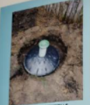 Round, dark-colored well access point, with a green cap and white pipe, in a dirt setting.