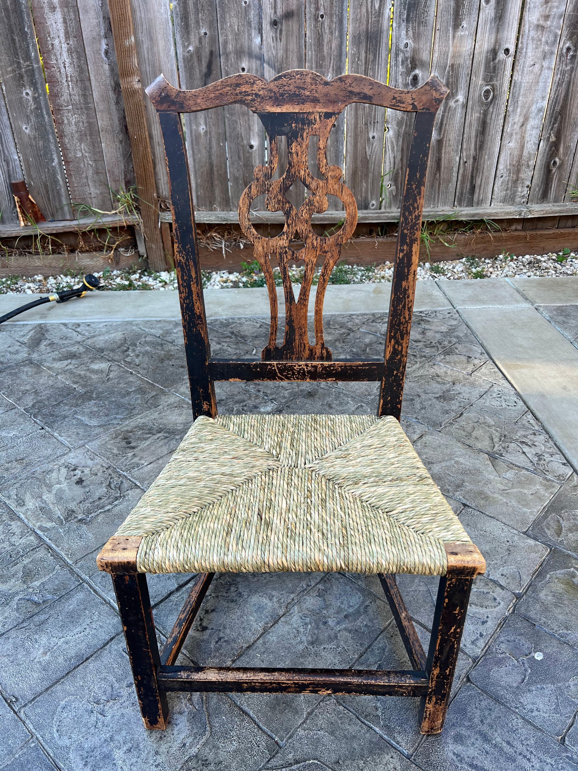 A wooden chair with a wicker seat is sitting on a sidewalk in front of a wooden fence.