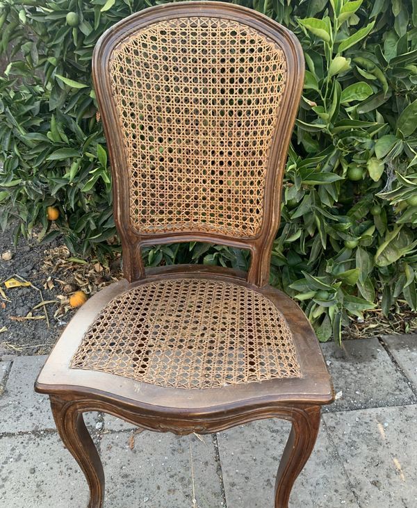 A wooden chair with a wicker seat is sitting on a sidewalk in front of a bush.