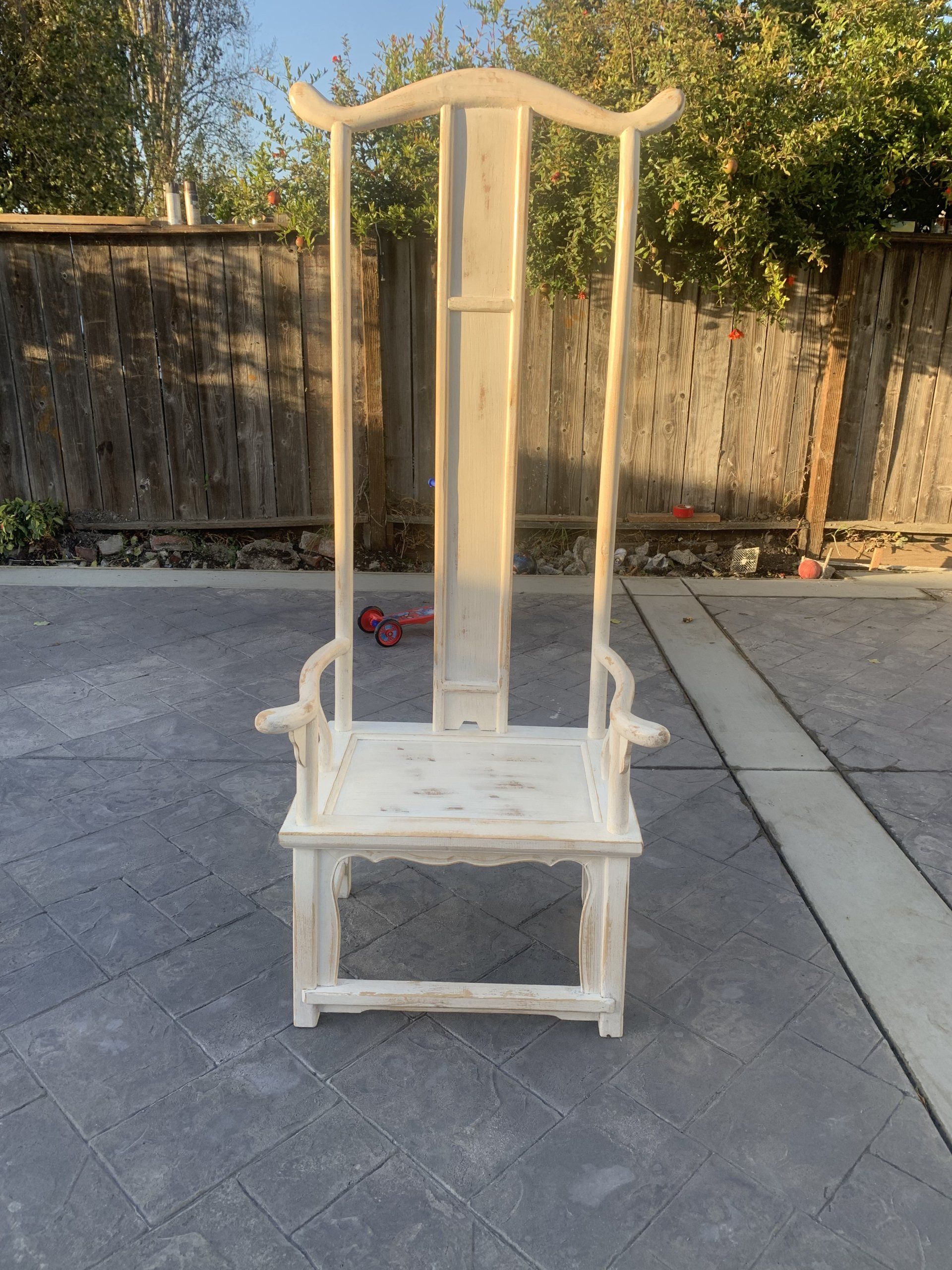 A white wooden chair is sitting on a concrete patio in front of a wooden fence.
