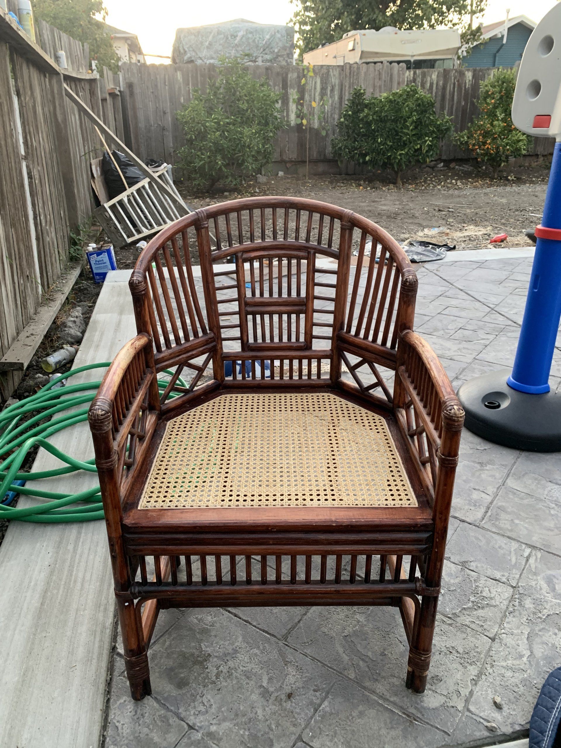 A wooden chair with a wicker seat is sitting on a patio next to a hose.