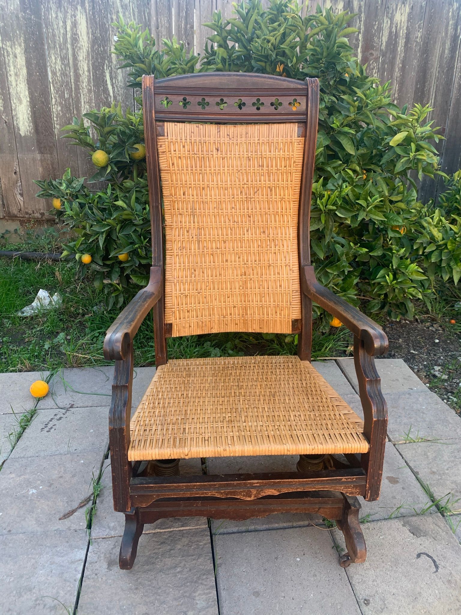 A wooden rocking chair with a wicker seat is sitting on a sidewalk in front of a bush.