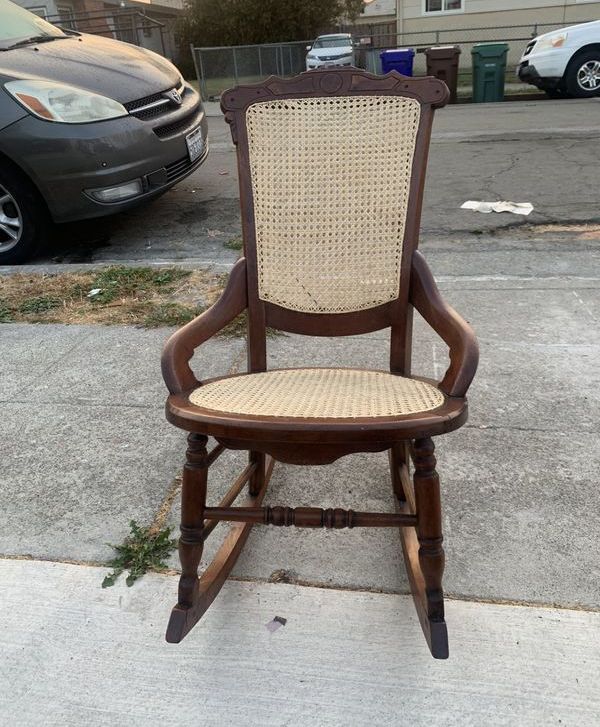 A wooden rocking chair with a wicker seat is parked on the sidewalk in front of a car.