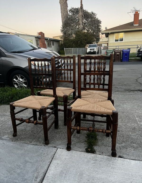 Three wooden chairs with wicker seats are sitting on a sidewalk in front of a car.