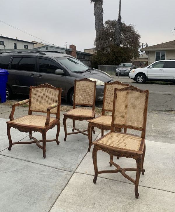 Four chairs with wicker seats are sitting on a sidewalk in front of a car.