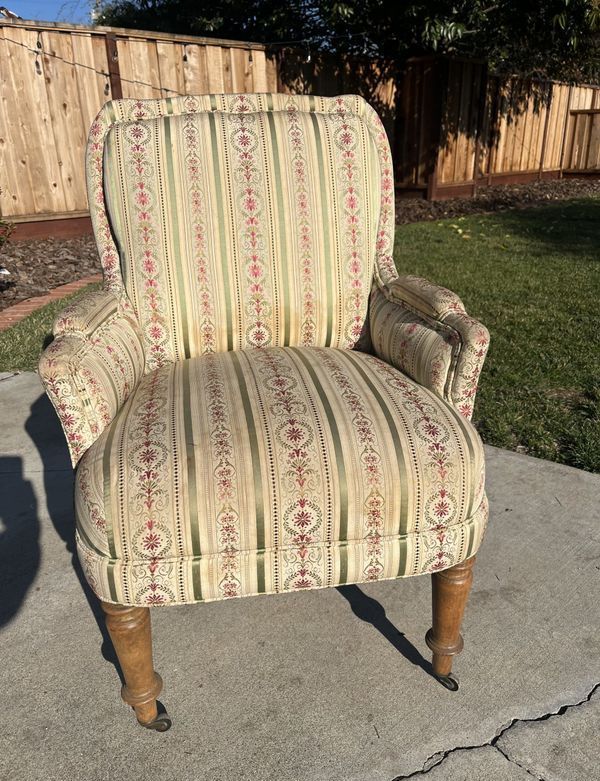 A chair with a striped fabric is sitting on a sidewalk in front of a wooden fence.