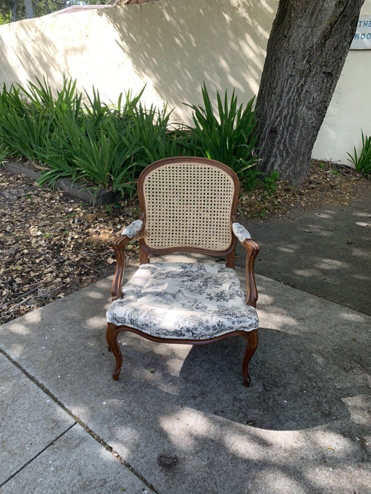 A chair with a cane back is sitting on a sidewalk next to a tree.