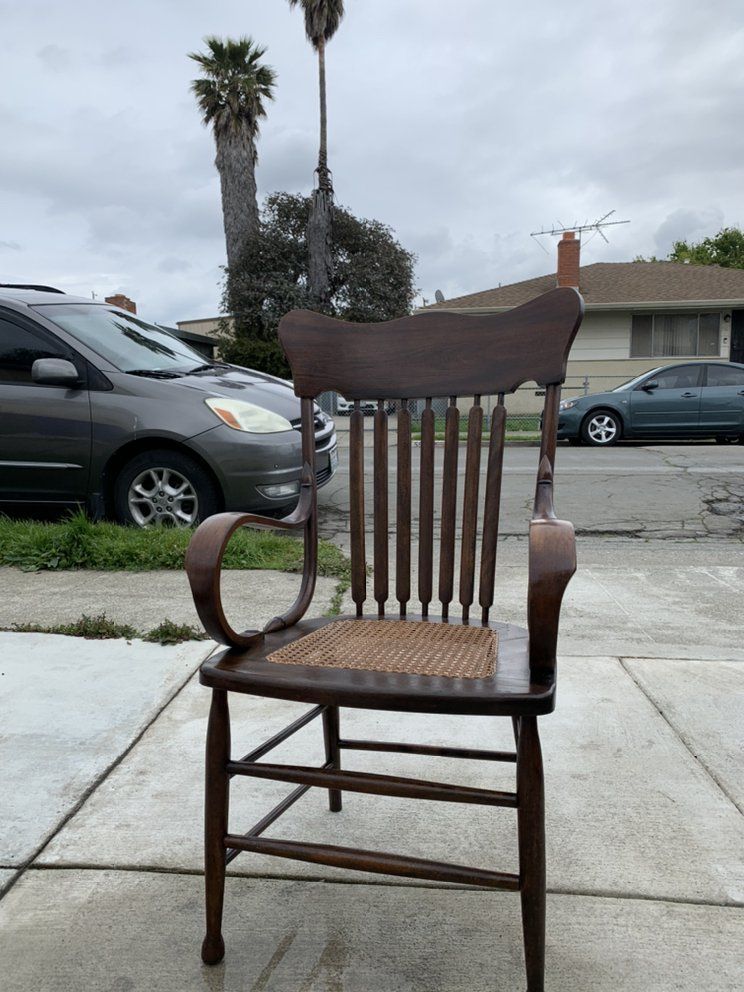 A wooden chair with a wicker seat is sitting on a sidewalk next to a car.