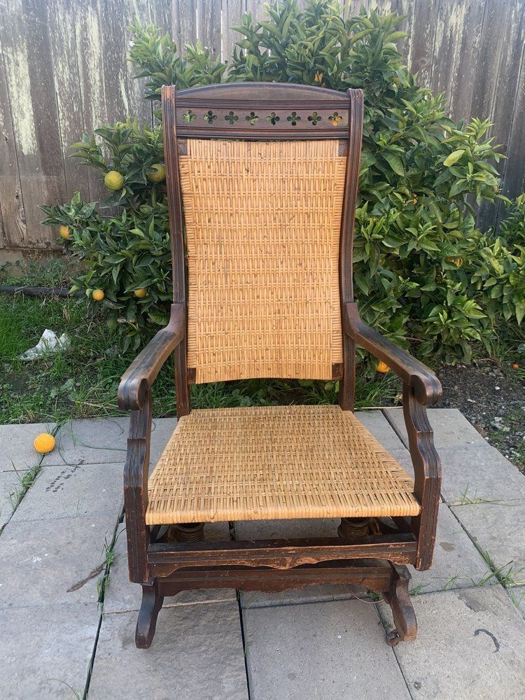 A wooden rocking chair with a wicker seat is sitting on a patio in front of a tree.