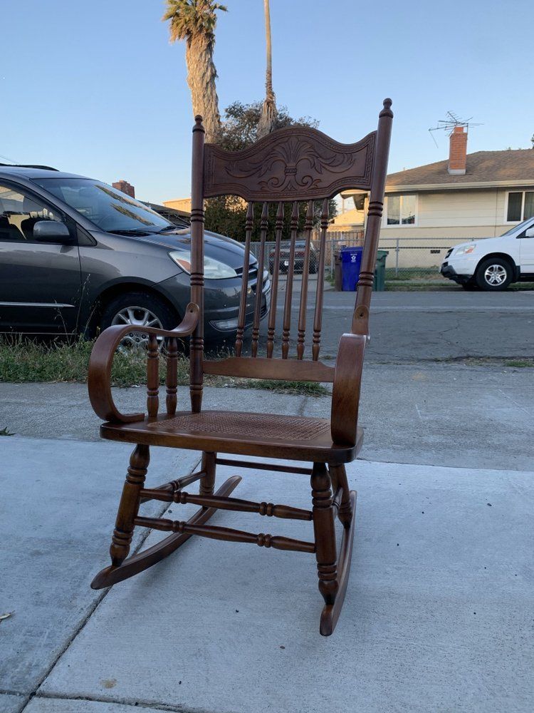 A wooden rocking chair is sitting on a sidewalk next to a car