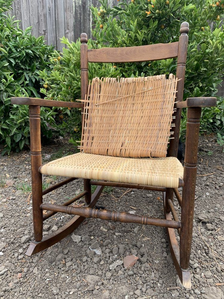 A wooden rocking chair with a wicker seat is sitting in the dirt in front of a bush.