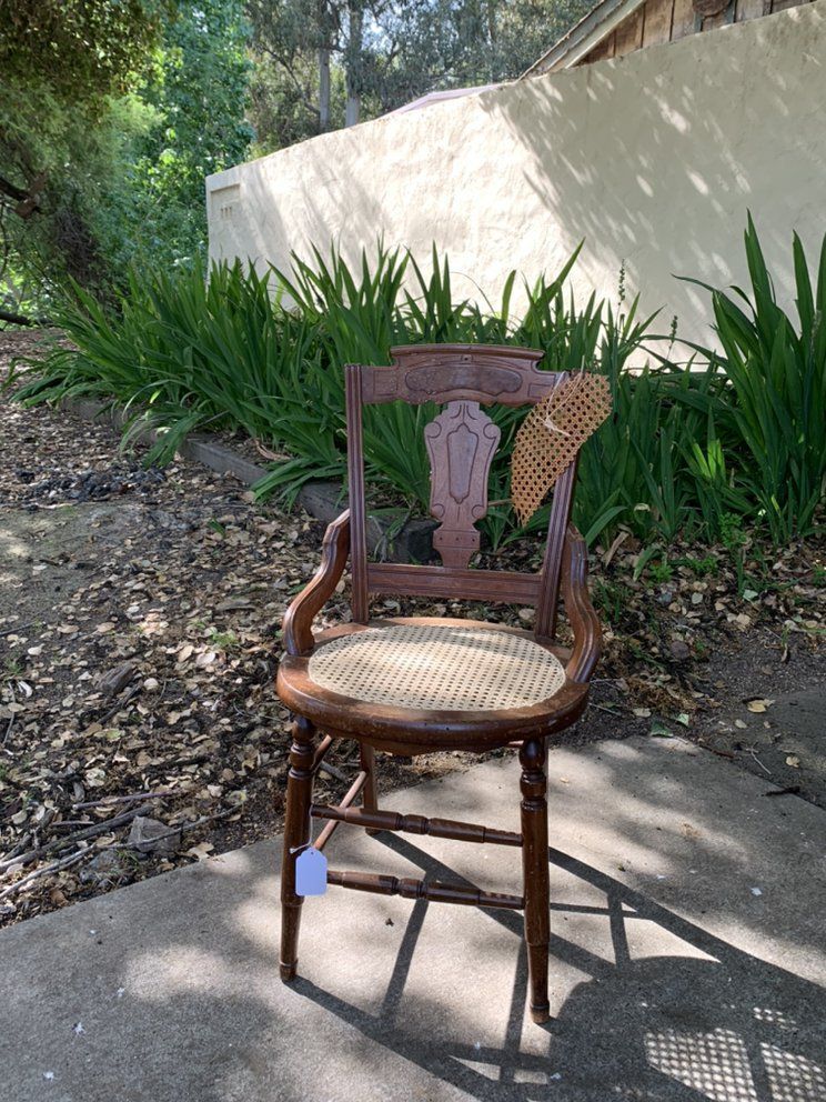 A wooden chair with a wicker seat is sitting on a sidewalk.