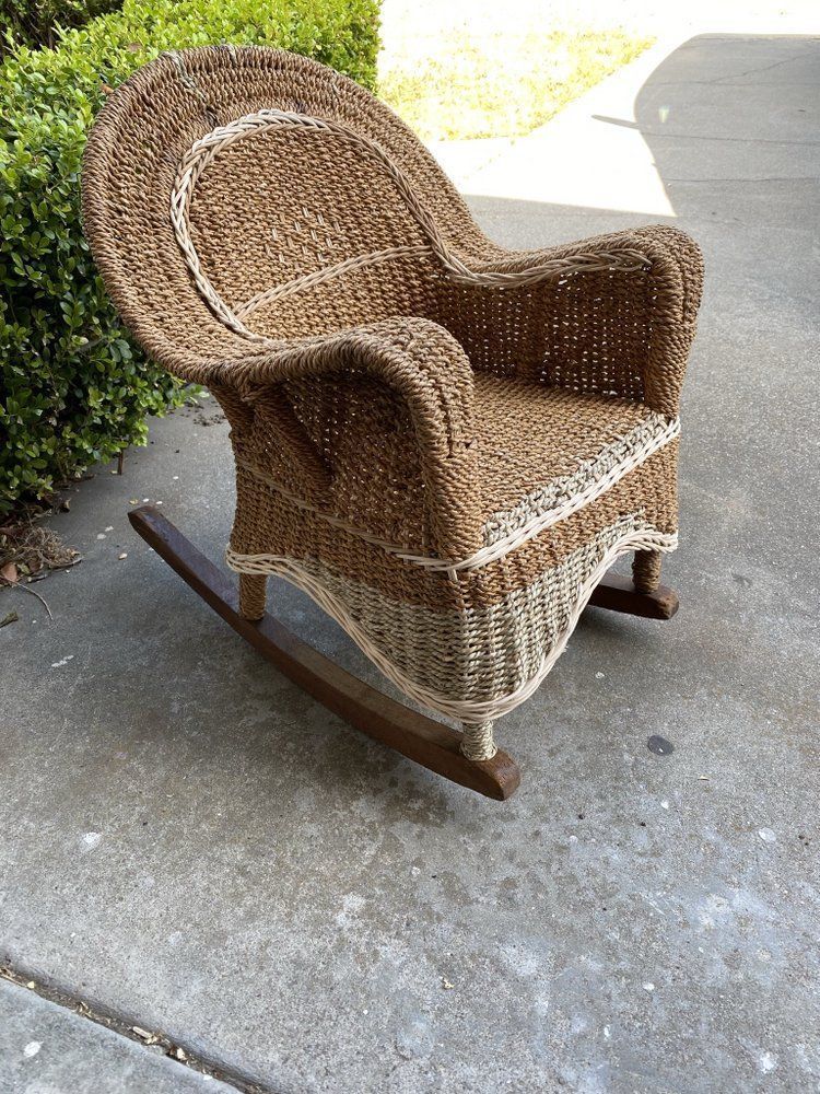 A wicker rocking chair is sitting on a sidewalk next to a bush.