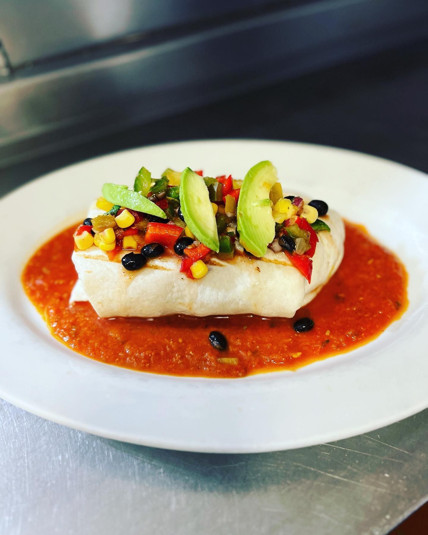 A white plate topped with a burrito covered in sauce and avocado slices.