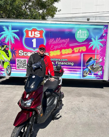 Person in red shirt stands with a red scooter in front of a US 1 Scooters advertisement on a trailer.
