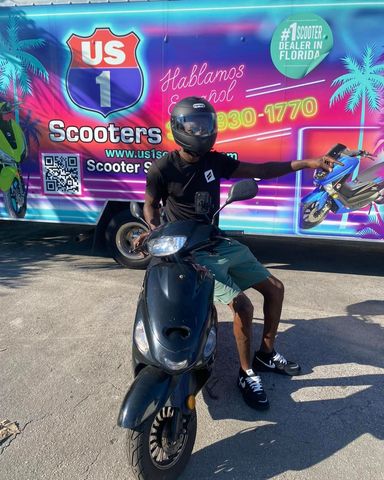 Person on black scooter in front of a US 1 Scooters vehicle; Florida setting.