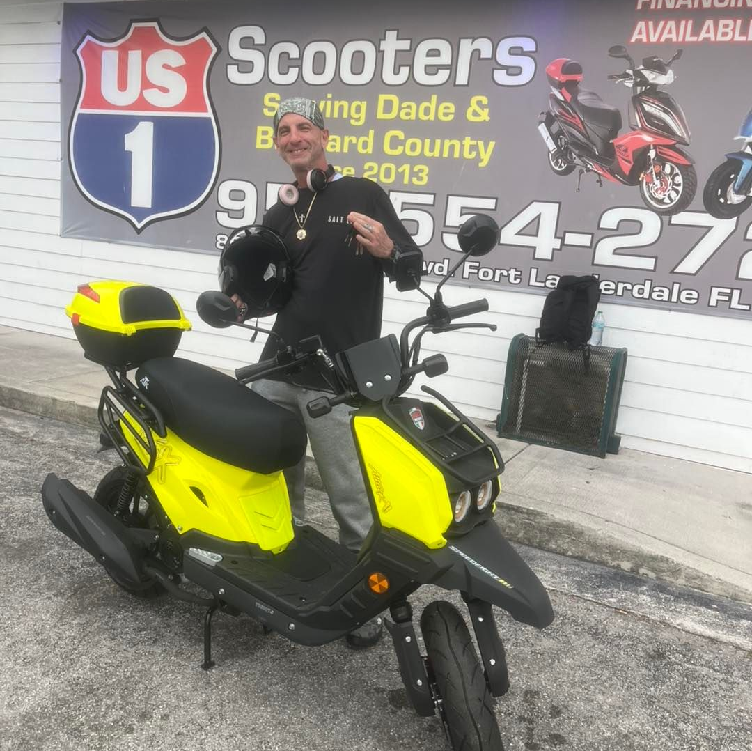 Man poses with a yellow scooter in front of a scooter shop.