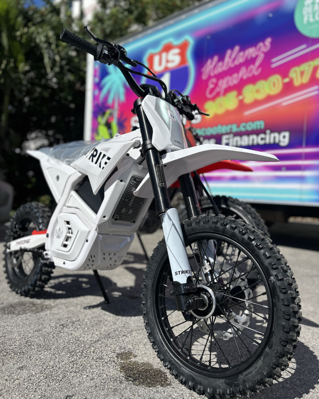White electric dirt bike parked outdoors with a blue and red sign in the background.