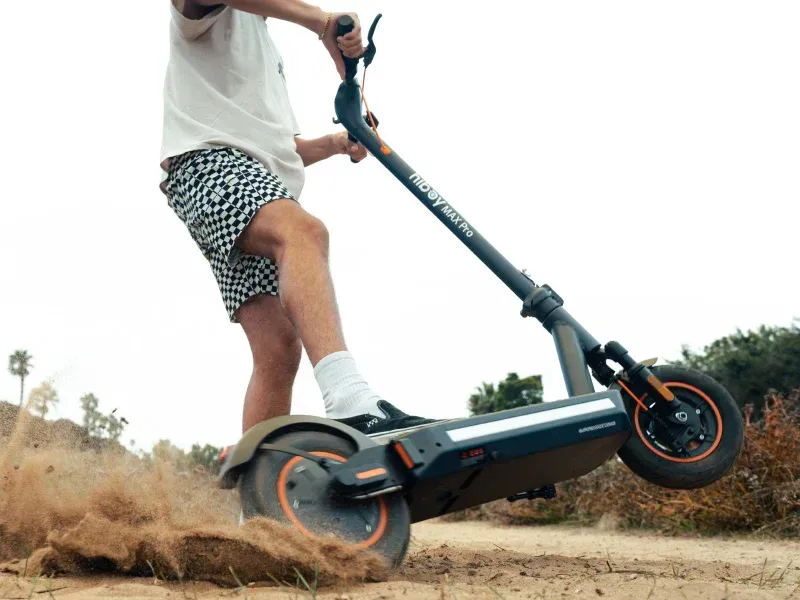 Person riding an electric scooter on a dirt path, kicking up dust. Orange tires, dark scooter frame.