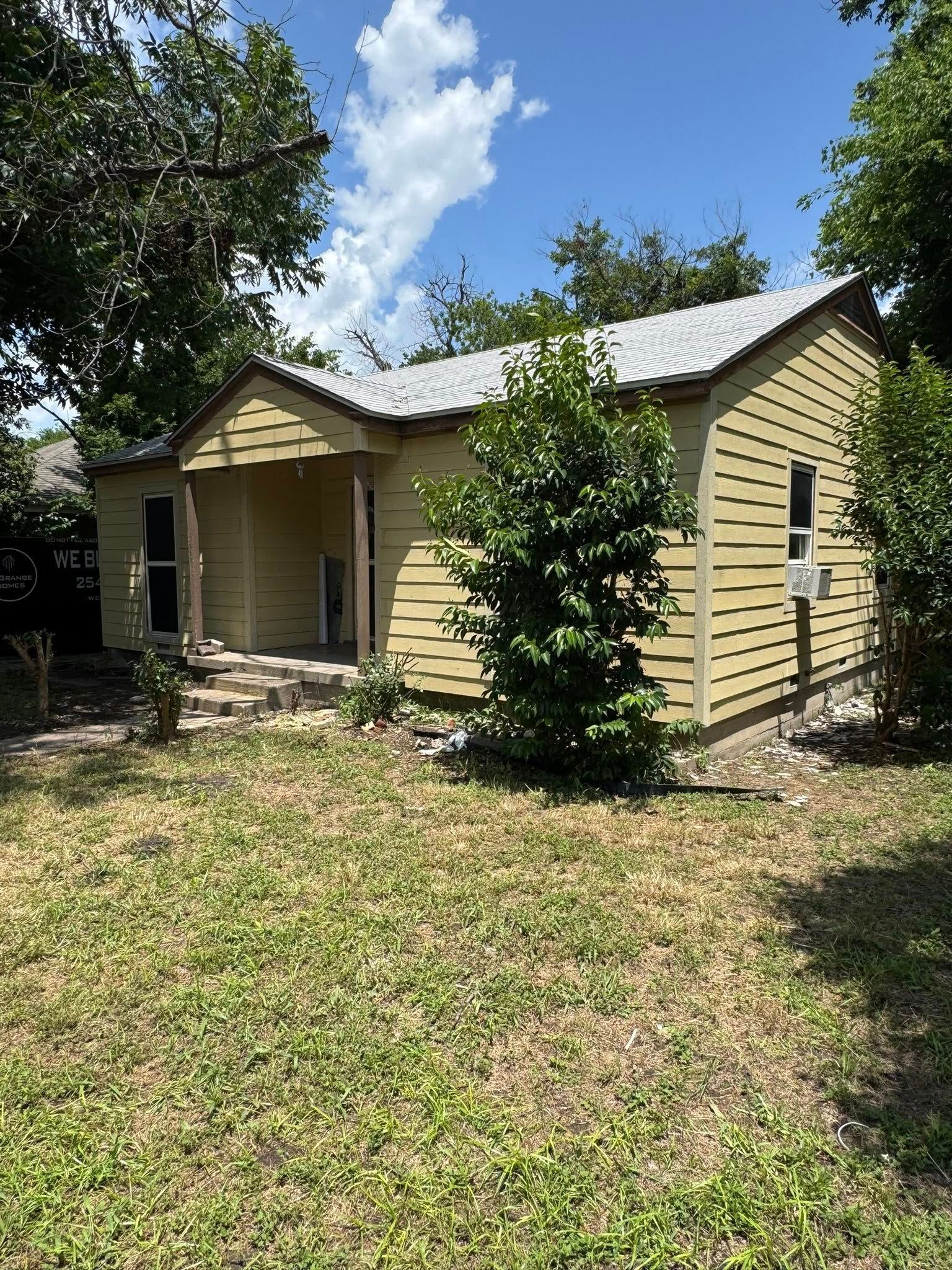 Yellow house with a small porch, surrounded by trees and grass, under a partly cloudy sky.