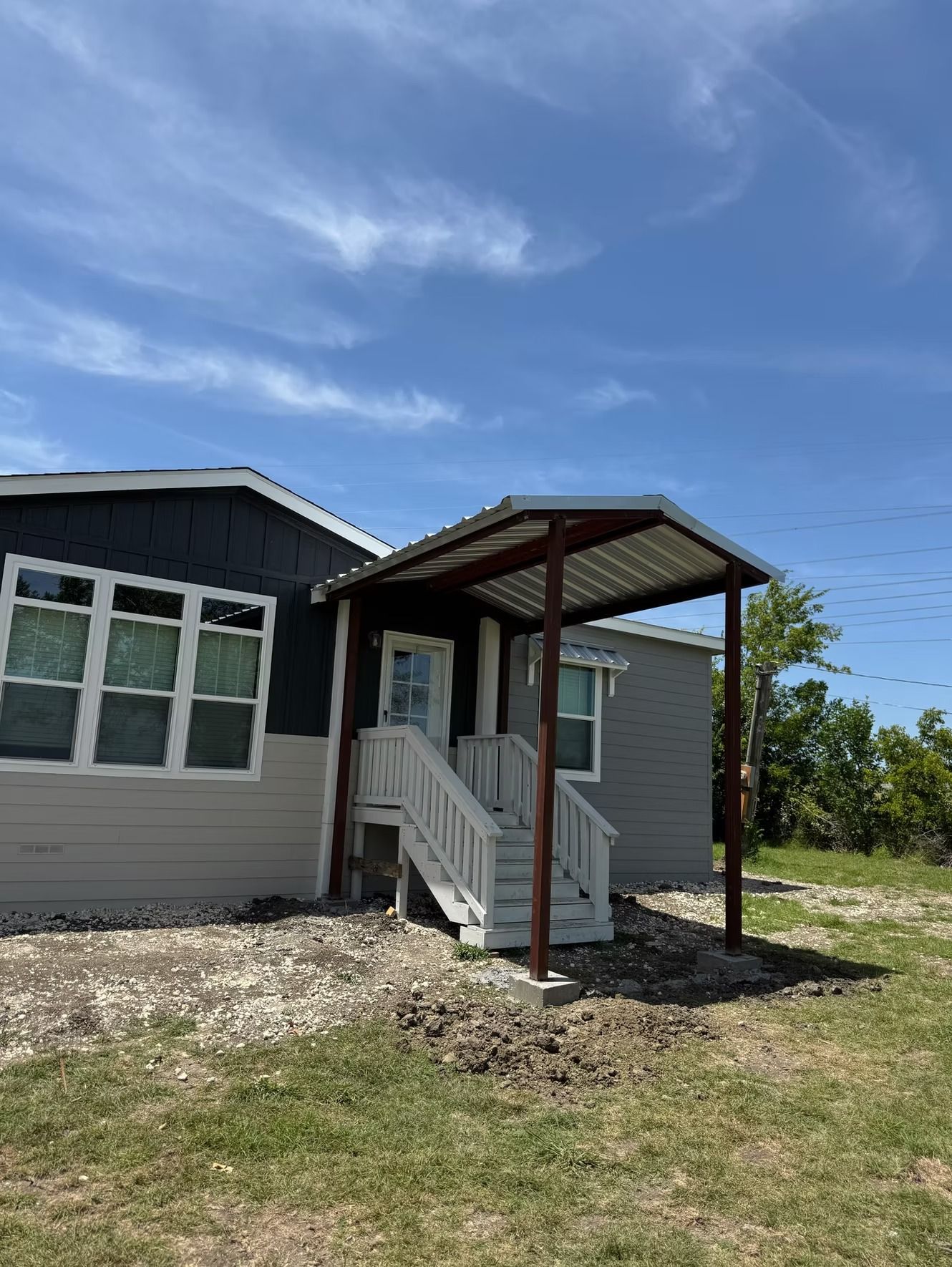 House exterior with a small porch, gray and blue siding, white stairs, and a metal roof under a blue sky.