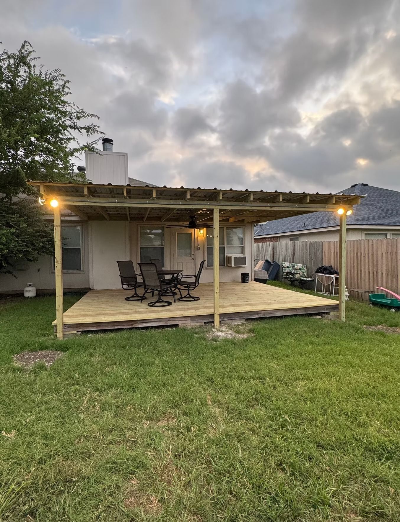 Wooden deck and pergola in a backyard, with a table, chairs, and cloudy sky.
