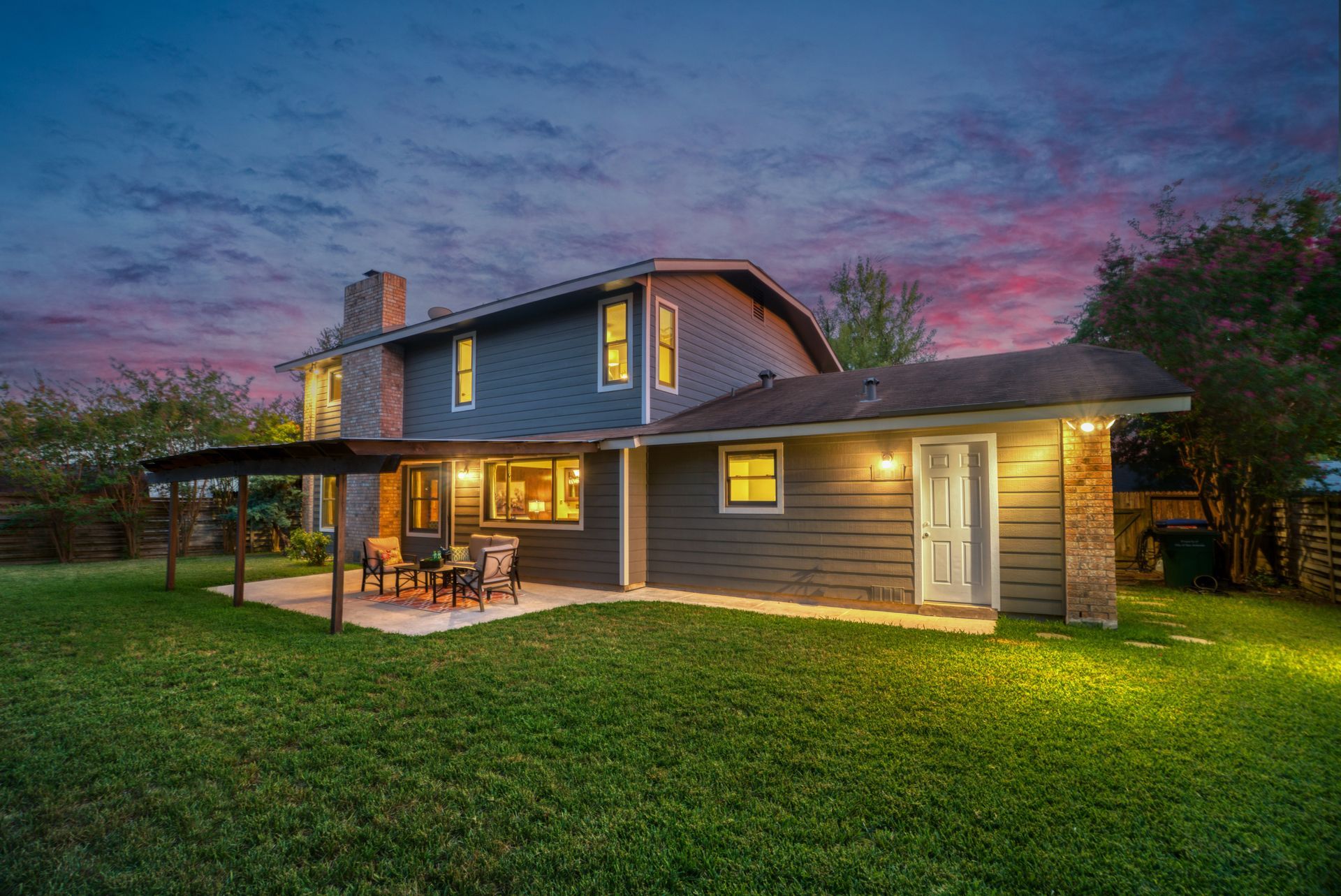 Rear view of a two-story blue house with an attached patio, lit up against a twilight sky.