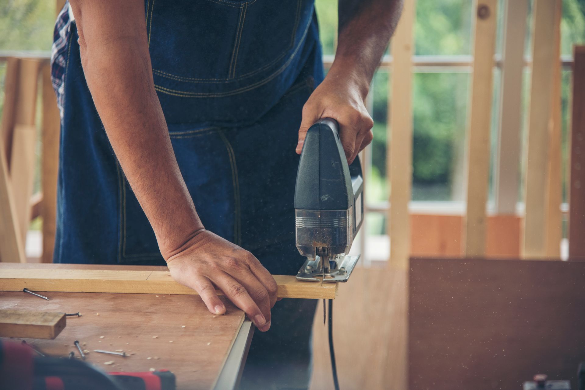 Person in blue apron using a jigsaw to cut a wooden plank on a workbench in a workshop.