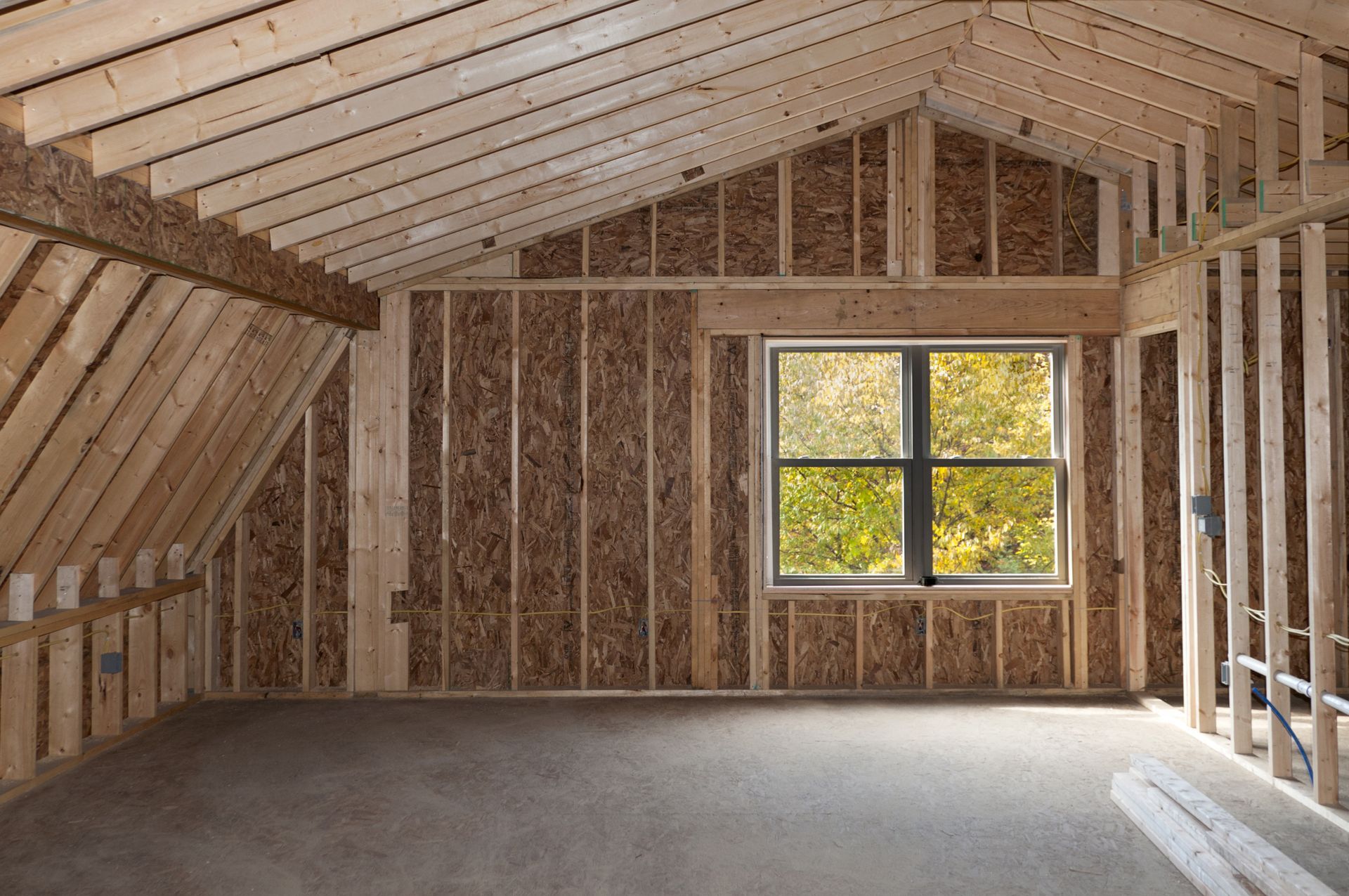 Interior of a room under construction, with wooden framing, sheathing, and a window overlooking trees.