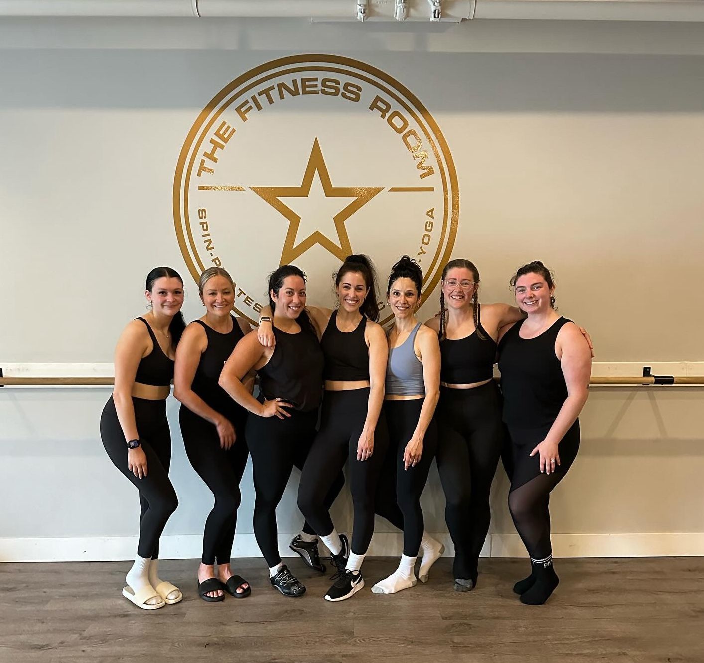 A group of women are posing for a picture in front of a wall that says the fitness room.