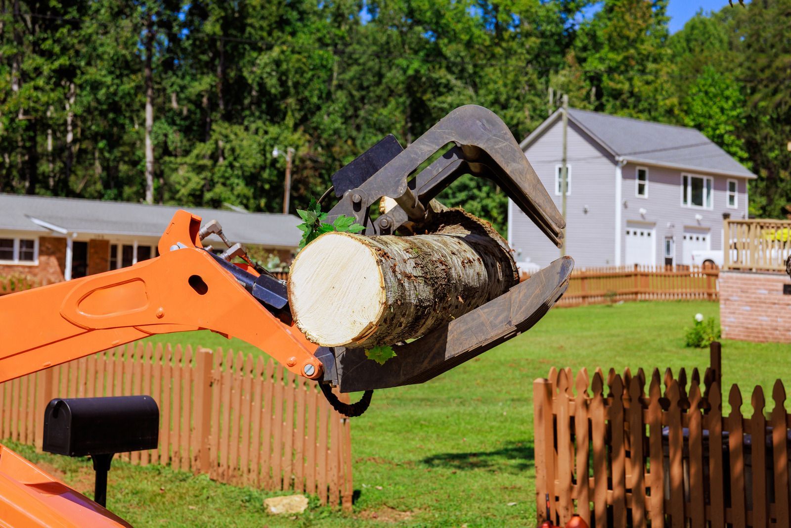 Orange excavator arm lifting a large cut tree trunk in a suburban yard