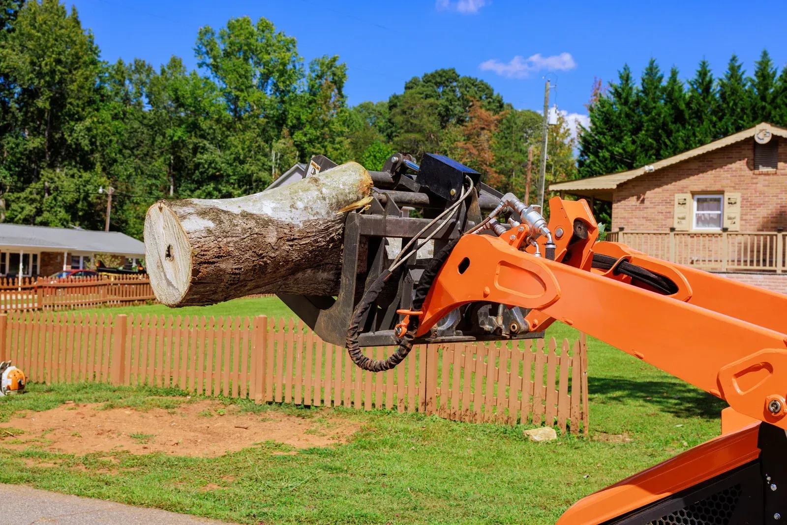 Orange excavator lifting a large tree trunk beside a house and wooden fence in a yard