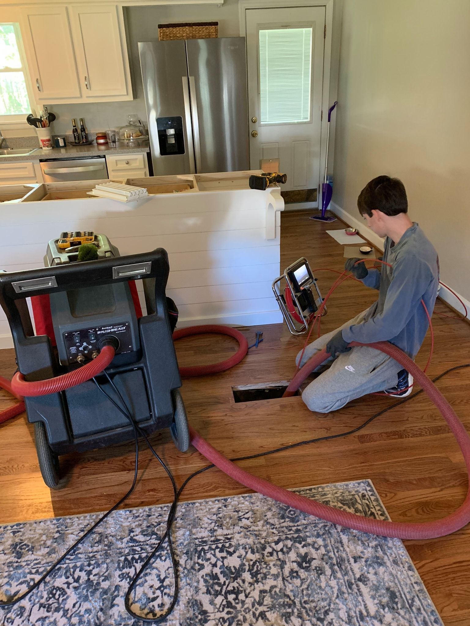 A person working on a floor in a kitchen. The worker is kneeling and wearing a grey shirt.