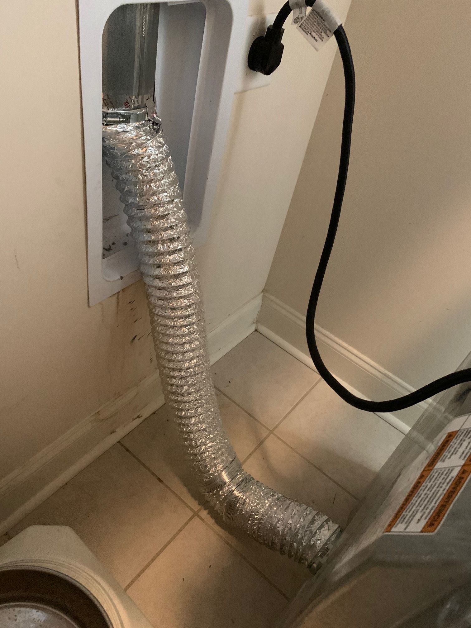 Dryer vent hose connected to a wall outlet. Silver hose bent downward. White wall and tile floor. Black power cord.