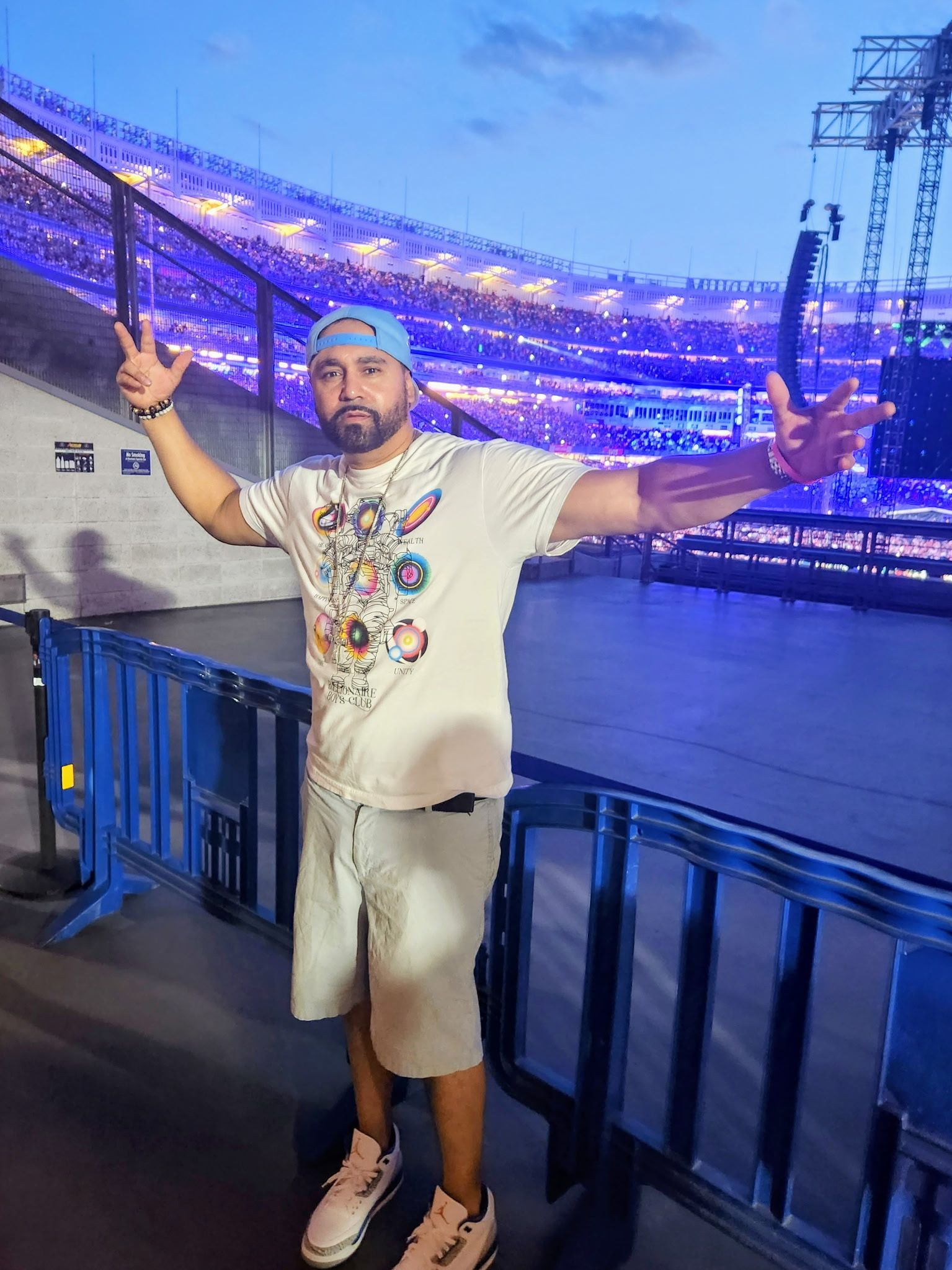 Man in a white shirt and shorts poses with arms out, stadium in the background lit in blue.