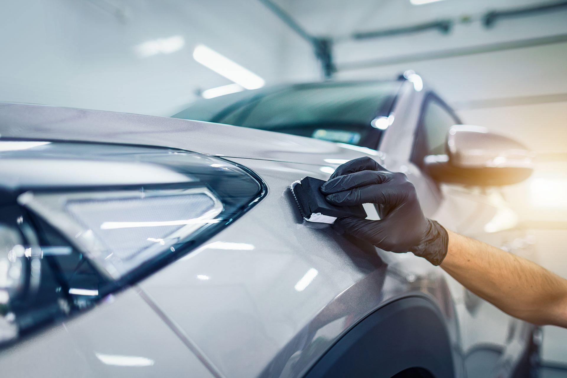 Person wearing black gloves polishing a silver car's front fender near the headlight inside a garage