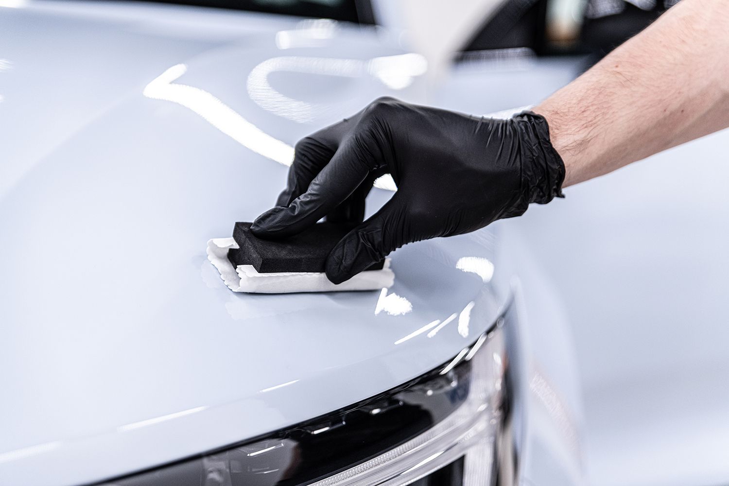 Close-up of ceramic coating being applied by a gloved detailer to a white car hood.