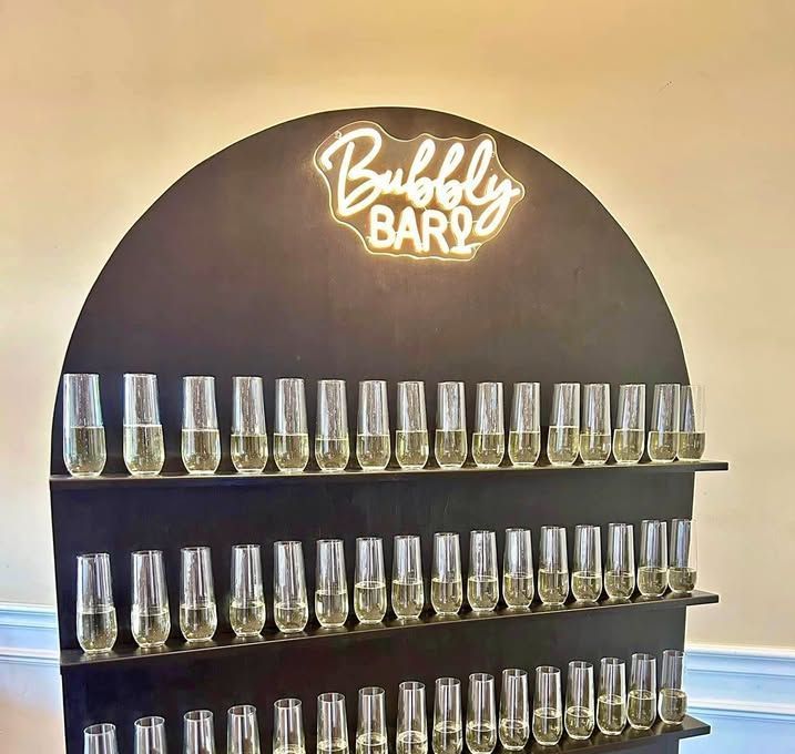 A display of wine glasses on a wall with a neon sign that says `` bubbly bar ''.