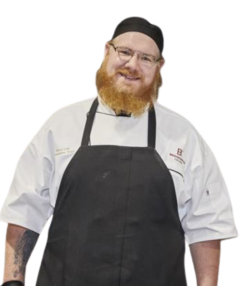 Chef in white coat, black apron and hat, smiling with red beard.