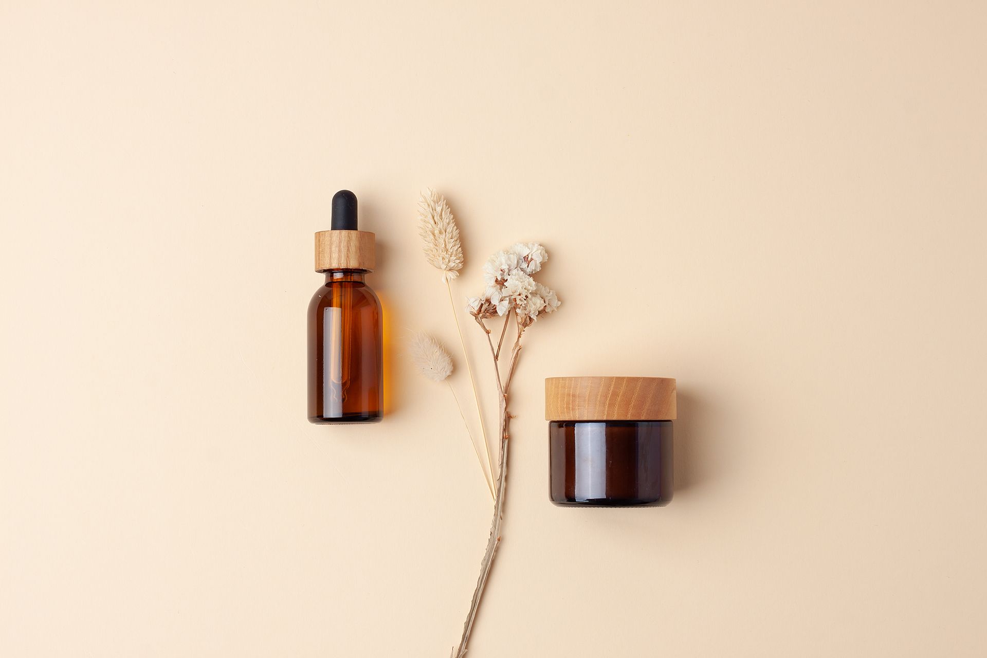 A bottle of essential oil , a jar of cream , and a dried flower on a beige background.