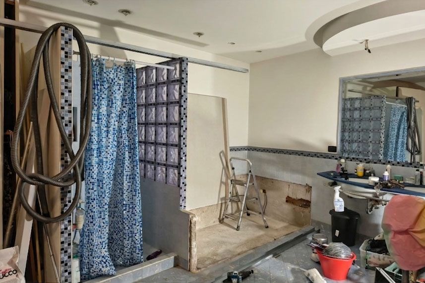 Un bagno in fase di ristrutturazione, con una parete doccia in blocchi di vetro, una tenda doccia con motivo blu e lavori in corso.