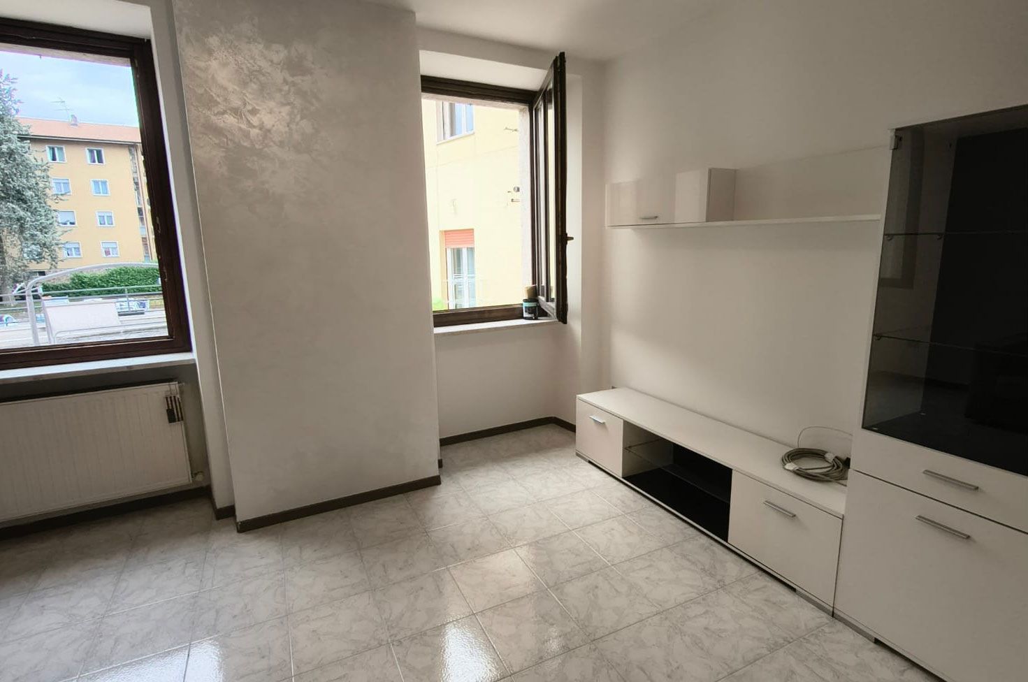 Una stanza luminosa e vuota con pavimento piastrellato, due finestre e armadietti bianchi lungo la parete.