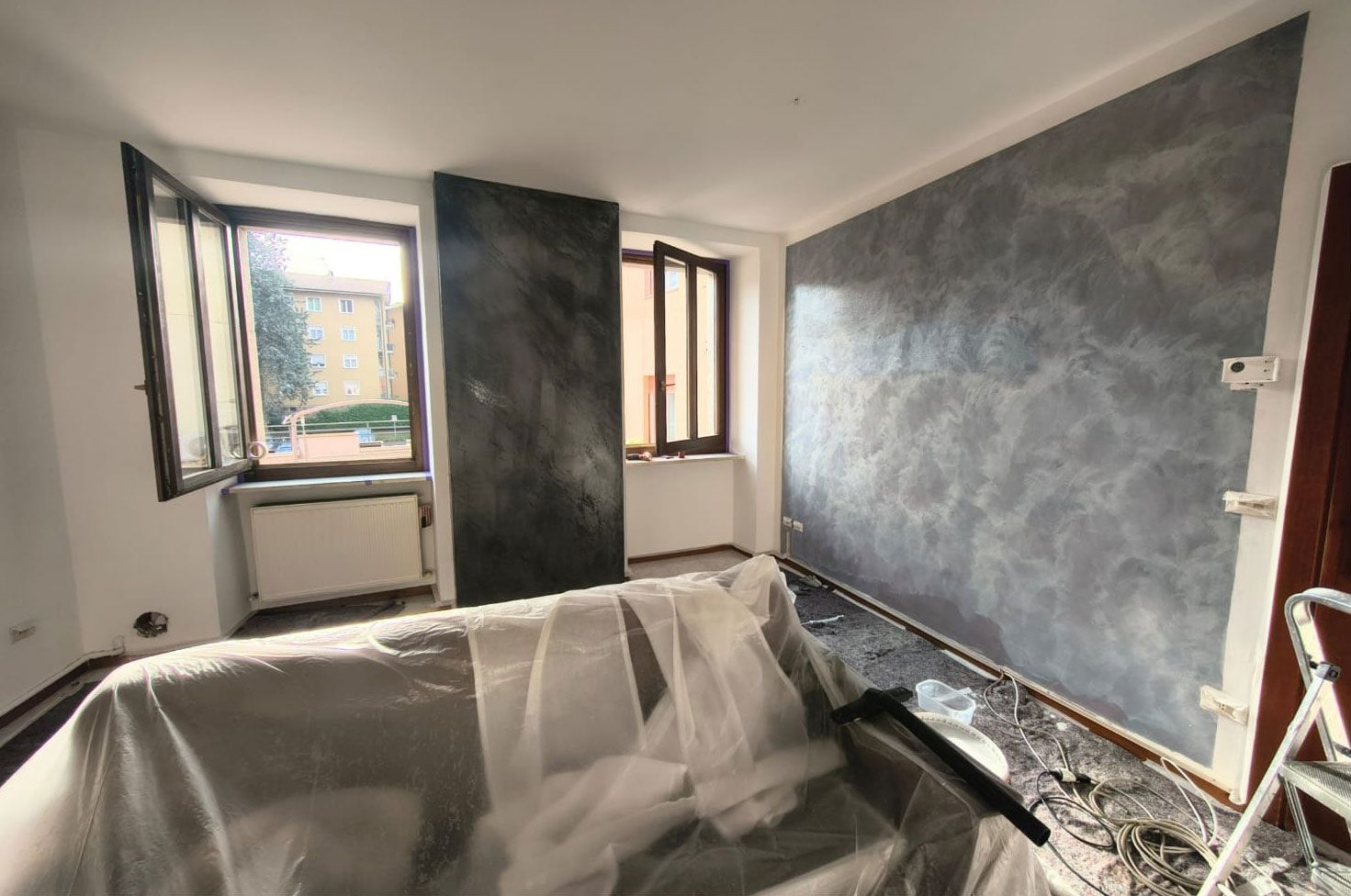Una stanza in fase di ristrutturazione con un'ampia parete decorativa grigia e texturizzata, una colonna centrale scura e un divano rivestito in plastica.