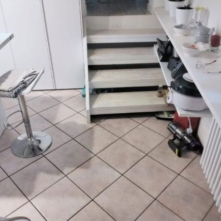 Una stanza con pavimento in piastrelle chiare, uno sgabello alto da bar, scale in legno e una mensola bianca con utensili da cucina.