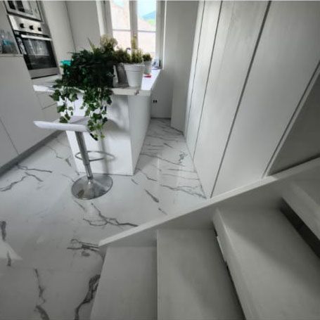 Una cucina moderna con pavimento effetto marmo, isola bianca, sgabello da bar e mobili bianchi, vista da una scala.