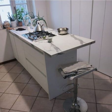 Una moderna isola cucina bianca con piano di lavoro in marmo, piano cottura a gas, piante in vaso e un singolo sgabello da bar con asciugamano.